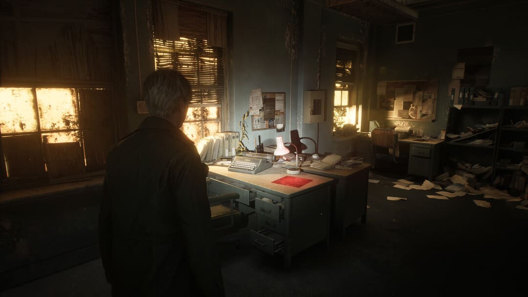 게임 Silent Hill 2 (2024)의 스크린 샷은 타자기와 느슨한 종이가있는 버려진 사무실 내부의 남자를 보여줍니다.